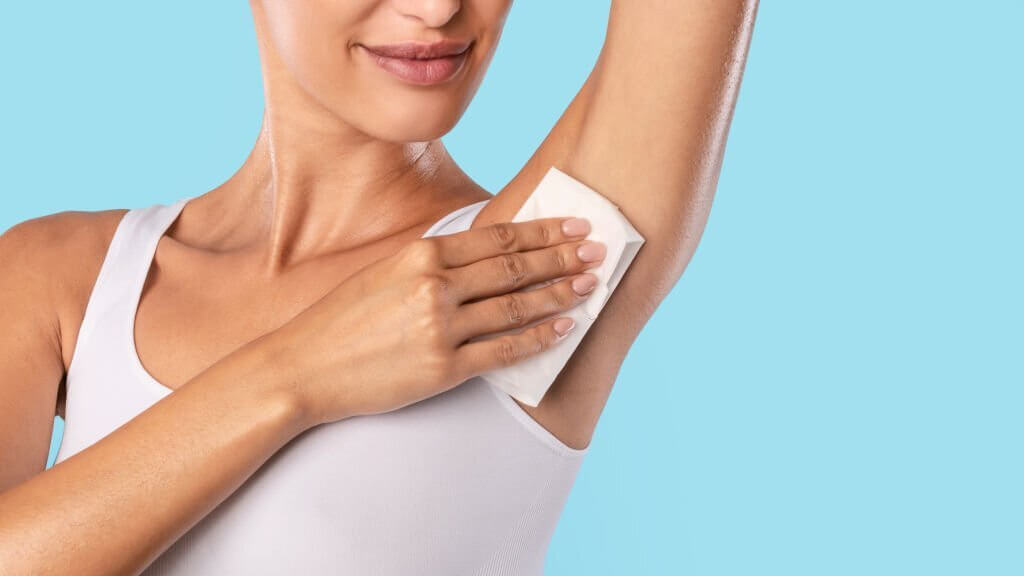 Closeup cropped view of young lady wiping her under arm area with napkin towel, rubbing armpit isolated on blue studuo background, putting hand up, touching axilla