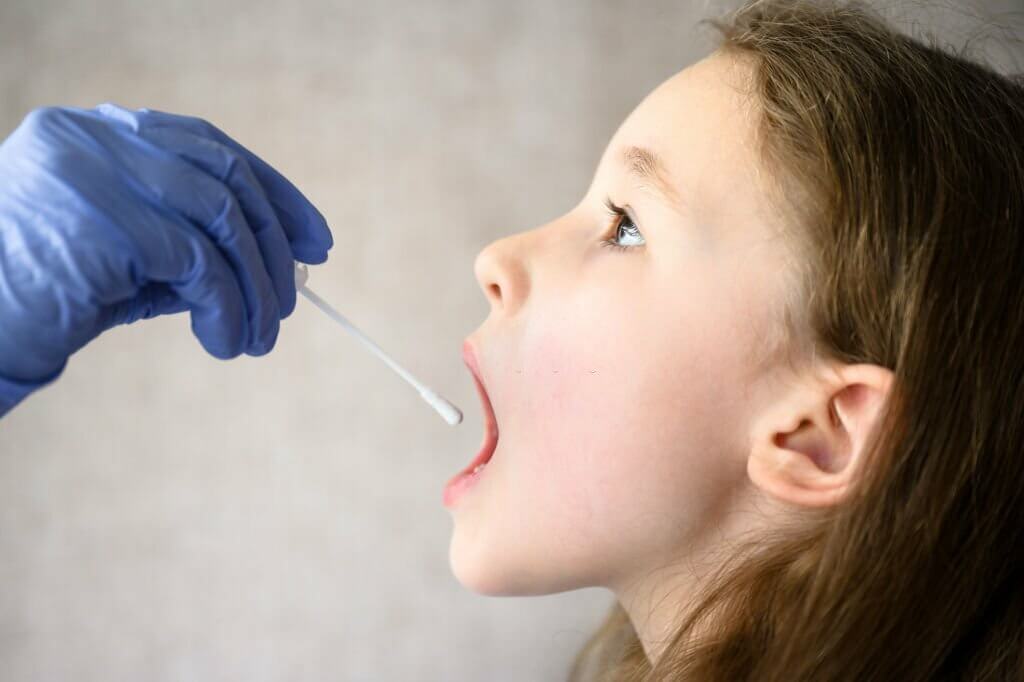 Child opens mouth for COVID-19 PCR test, doctor holds swab for saliva sample from cute kid during coronavirus pandemic.