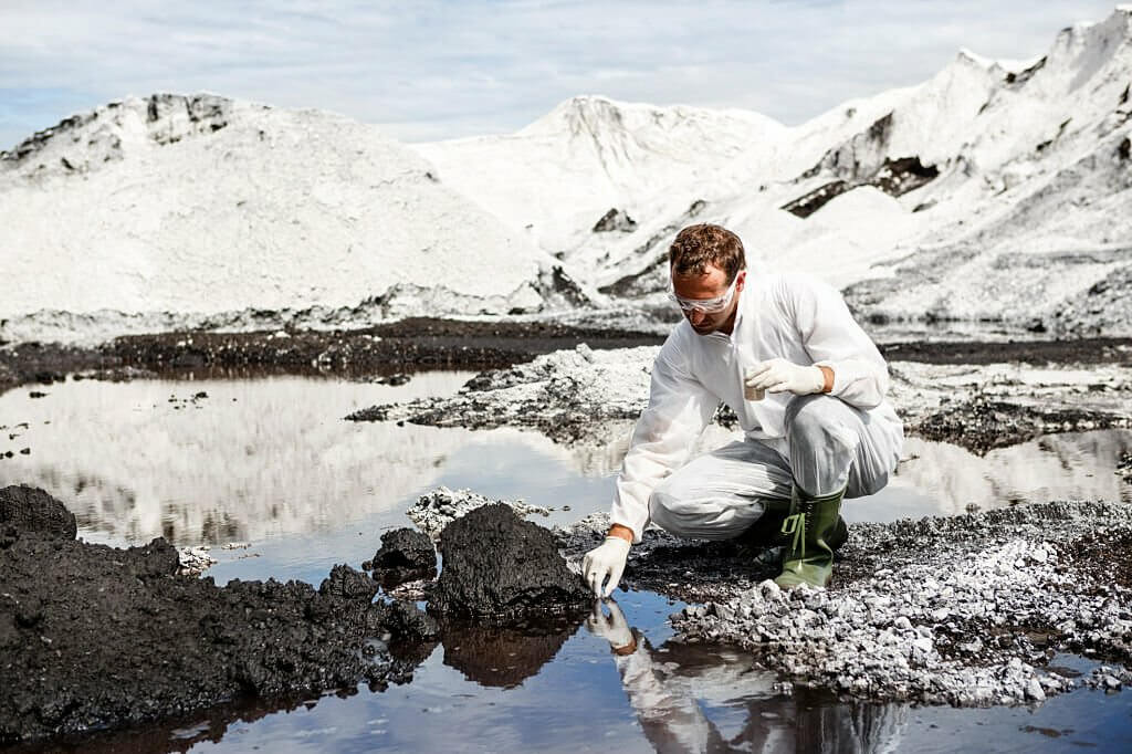 Scientist in white safety suit examing polluted water in a river at industrial site.